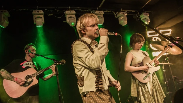 Trzech muzyków na scenie podczas koncertu, wokalista w centrum śpiewa do mikrofonu, z lewej gitarzysta akustyczny, z prawej gitarzystka elektryczna, tło oświetlone zielonym światłem scenicznym.