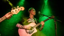 Mężczyzna z tatuażami na rękach gra na gitarze akustycznej i śpiewa do mikrofonu podczas koncertu na scenie oświetlonej zielonym światłem, w tle widoczny fragment gitary basowej drugiego muzyka.