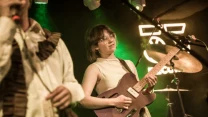 kobieta grająca na gitarze elektrycznej na scenie, w tle widoczne bębny oraz światła sceniczne w odcieniach zieleni, obok niej stoi osoba ubrana w biały strój z brązowymi elementami