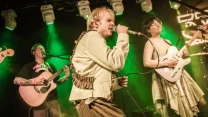 Zespół muzyczny występuje na scenie w świetle zielonych reflektorów; wokalista śpiewa do mikrofonu, obok niego gitarzystka gra na elektrycznej gitarze, a kolejny członek zespołu akompaniuje na gitarze akustycznej.