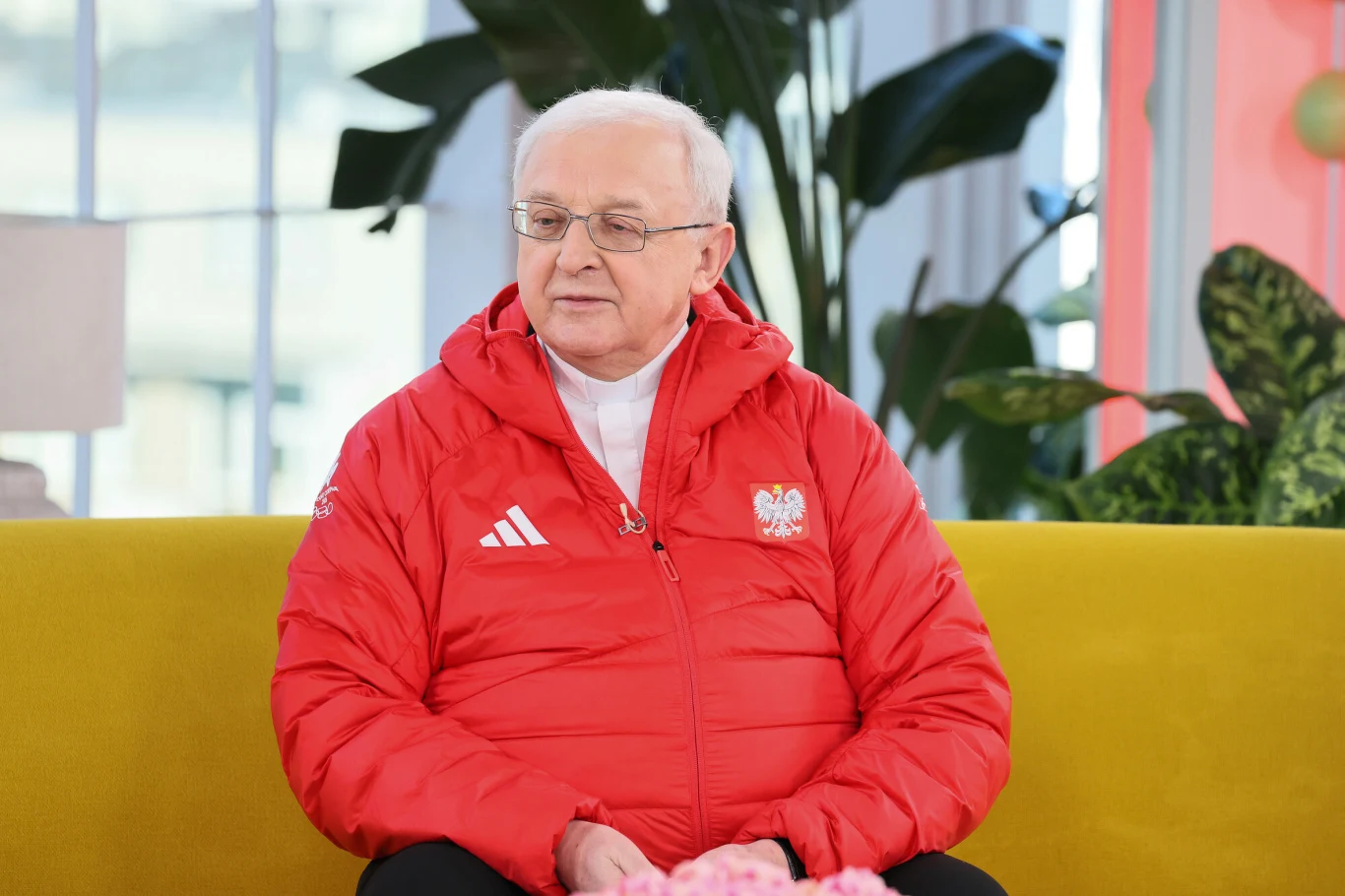 Ksiądz Edward Pleń starszy mężczyzna w okularach siedzący na żółtej sofie, ubrany w czerwoną kurtkę z godłem Polski i logo Adidas, w tle widoczne rośliny doniczkowe oraz jasne wnętrze.