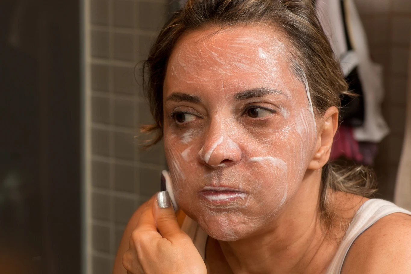 kobieta nakładająca krem lub maseczkę pielęgnacyjną na twarz, patrząca w lustro, w tle widoczna ściana wyłożona kafelkami