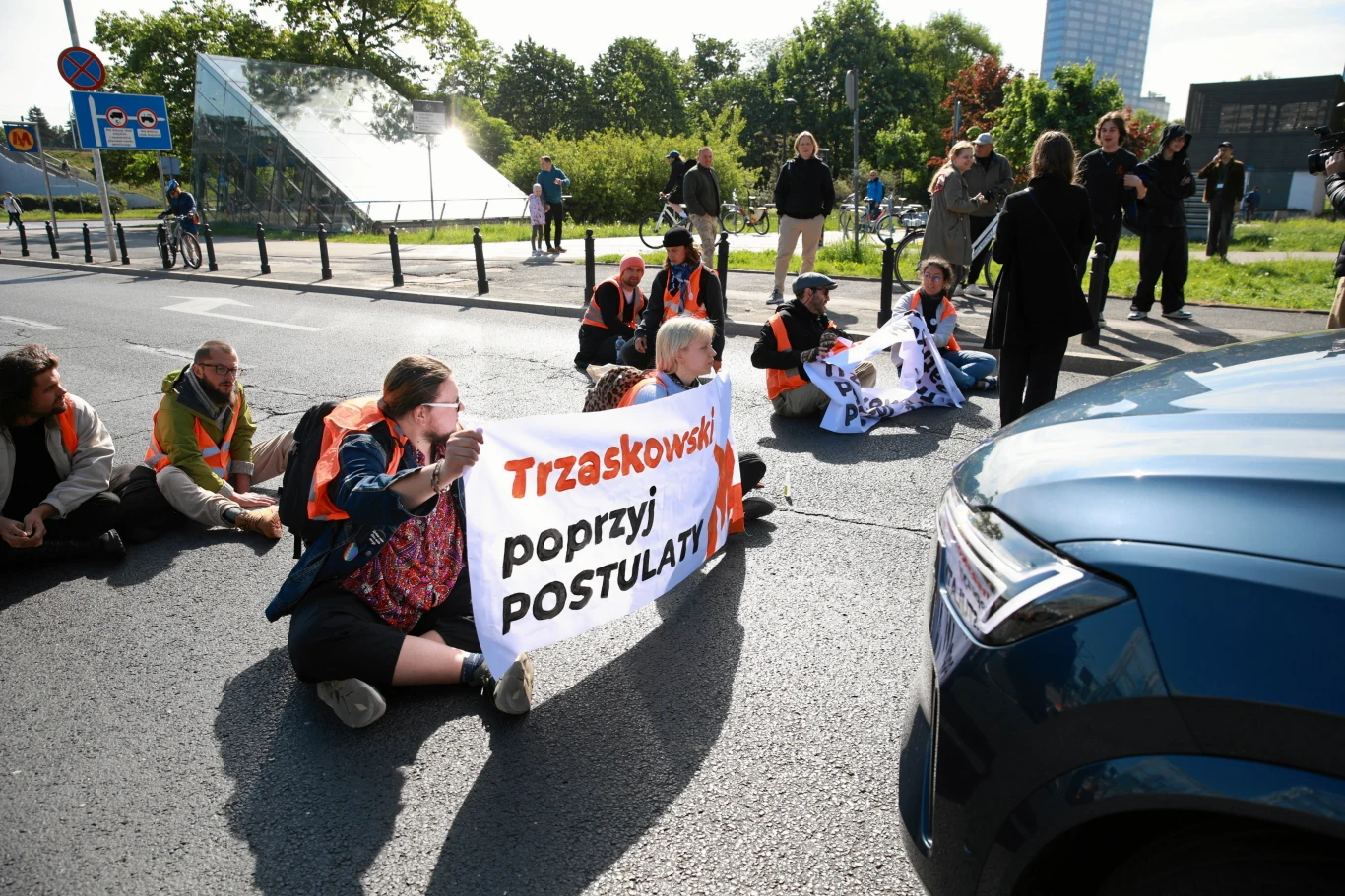 Grupa osób w pomarańczowych kamizelkach siedzi na jezdni, blokując ruch przed niebieskim samochodem, trzymając transparent z napisem 'Trzaskowski poprzyj postulaty'; w tle kilka osób stojących oraz zieleń miejska.