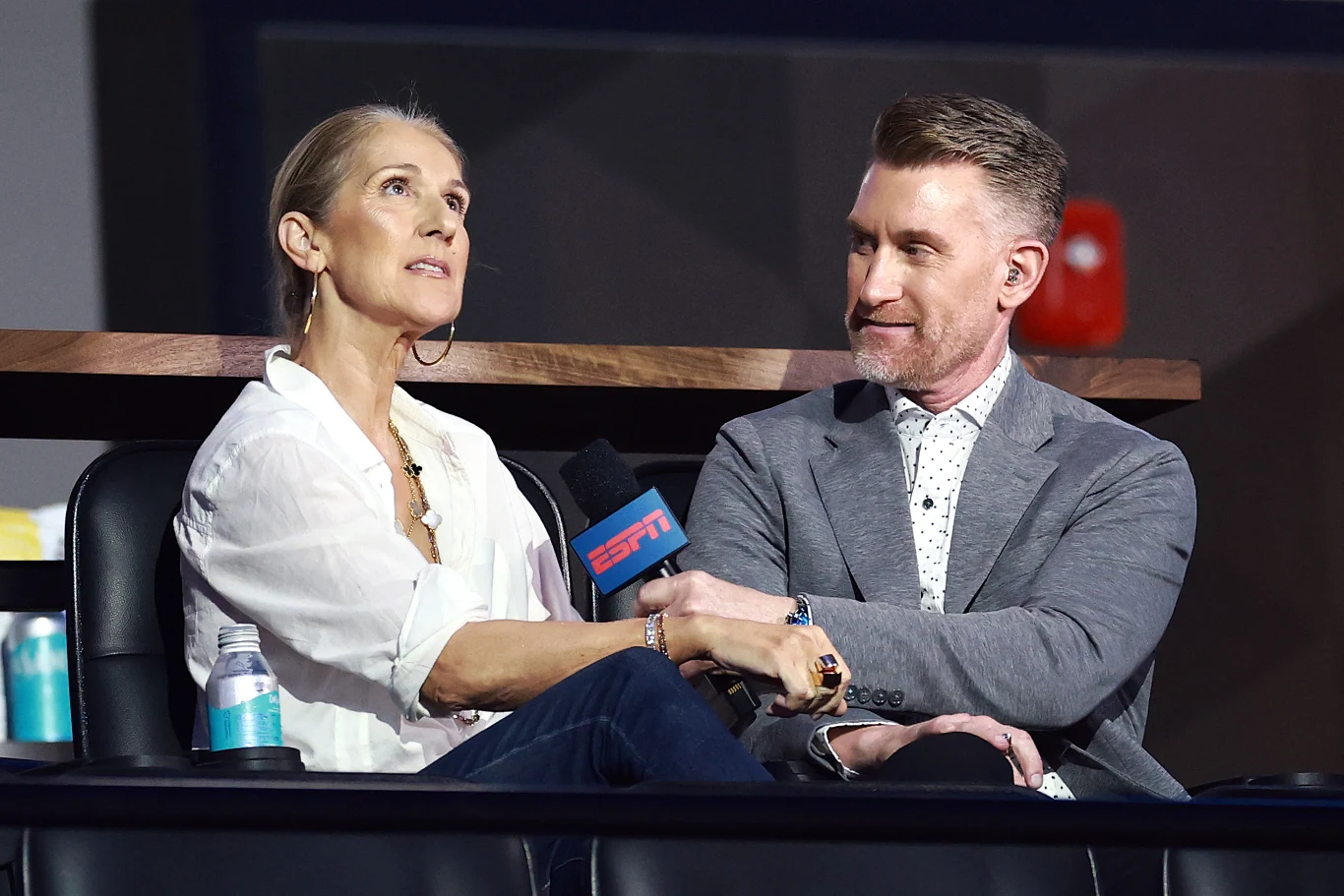 Céline Dion prezentuje nową piosenkę Céline Dion w białej koszuli i mężczyzna w szarej marynarce podczas wywiadu z mikrofonem ESPN, na stoliku butelki wody.