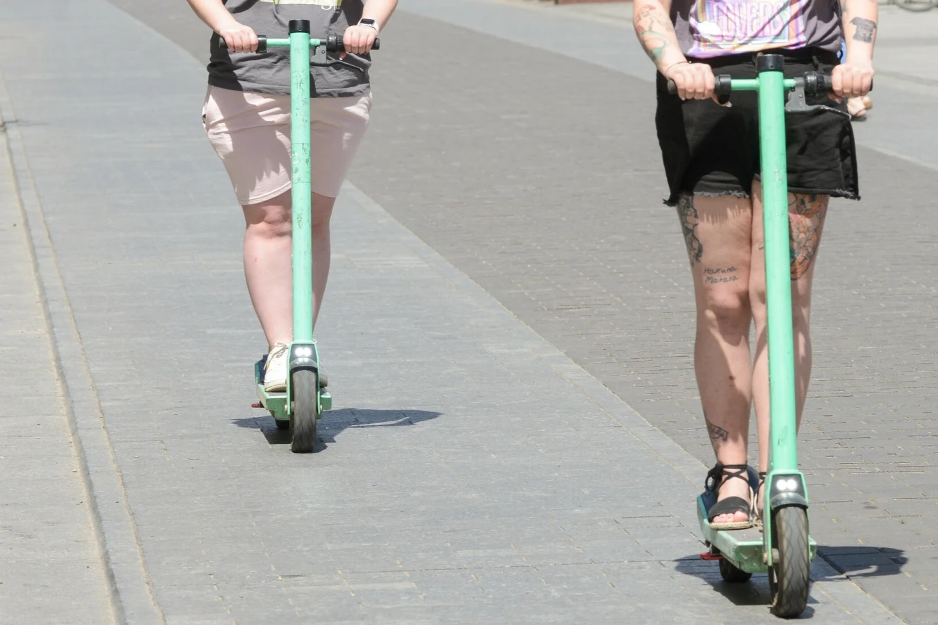 Hulajnogi elektryczne są bardziej niebezpieczne niż motocykle. Dwie osoby jadą na zielonych hulajnogach elektrycznych po chodniku; jedna ma tatuaże na nogach, druga jasną spódnicę.