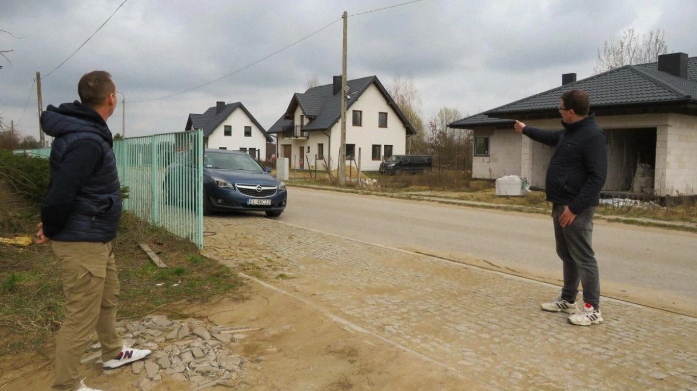 Dwóch mężczyzn stojących po przeciwnych stronach ulicy, jeden pokazuje ręką w kierunku pustostanu w trakcie budowy, po ulicy jedzie ciemny samochód, w tle widać nowe, białe domy jednorodzinne pod pochmurnym niebem.