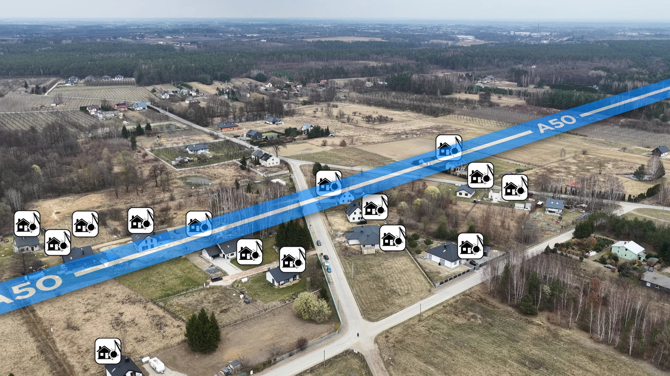 Wiejska okolica z rozproszoną zabudową jednorodzinną, przez którą przebiega wizualizacja szerokiej trasy oznaczonej jako A50; na trasie i wokół niej zaznaczono lokalizacje maszyn budowlanych, sugerujące planowaną budowę drogi i ingerencję w istniejące ...