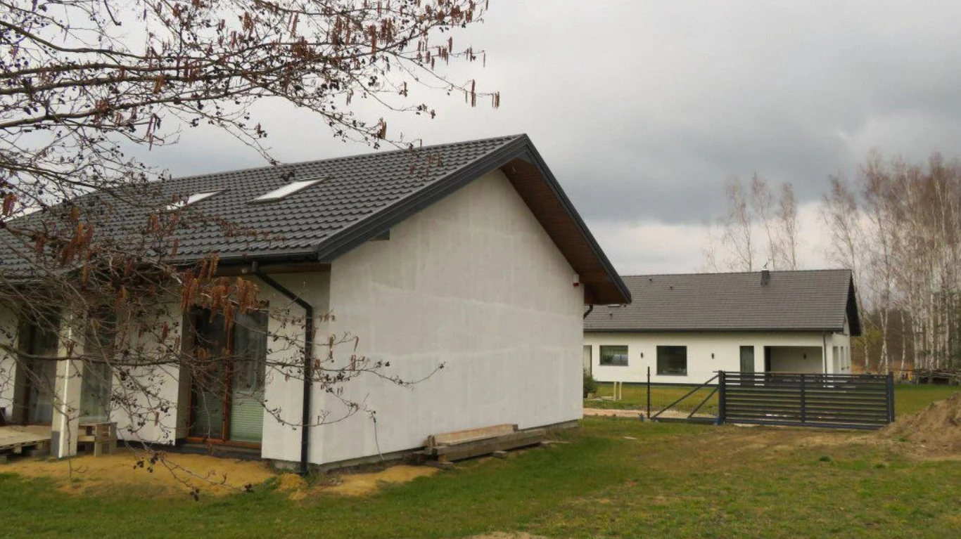 Dwa nowoczesne, parterowe domy o jasnych elewacjach i ciemnych dachach stoją na ogrodzonej działce, otoczone trawą i nielicznymi drzewami bez liści, pod zachmurzonym niebem.