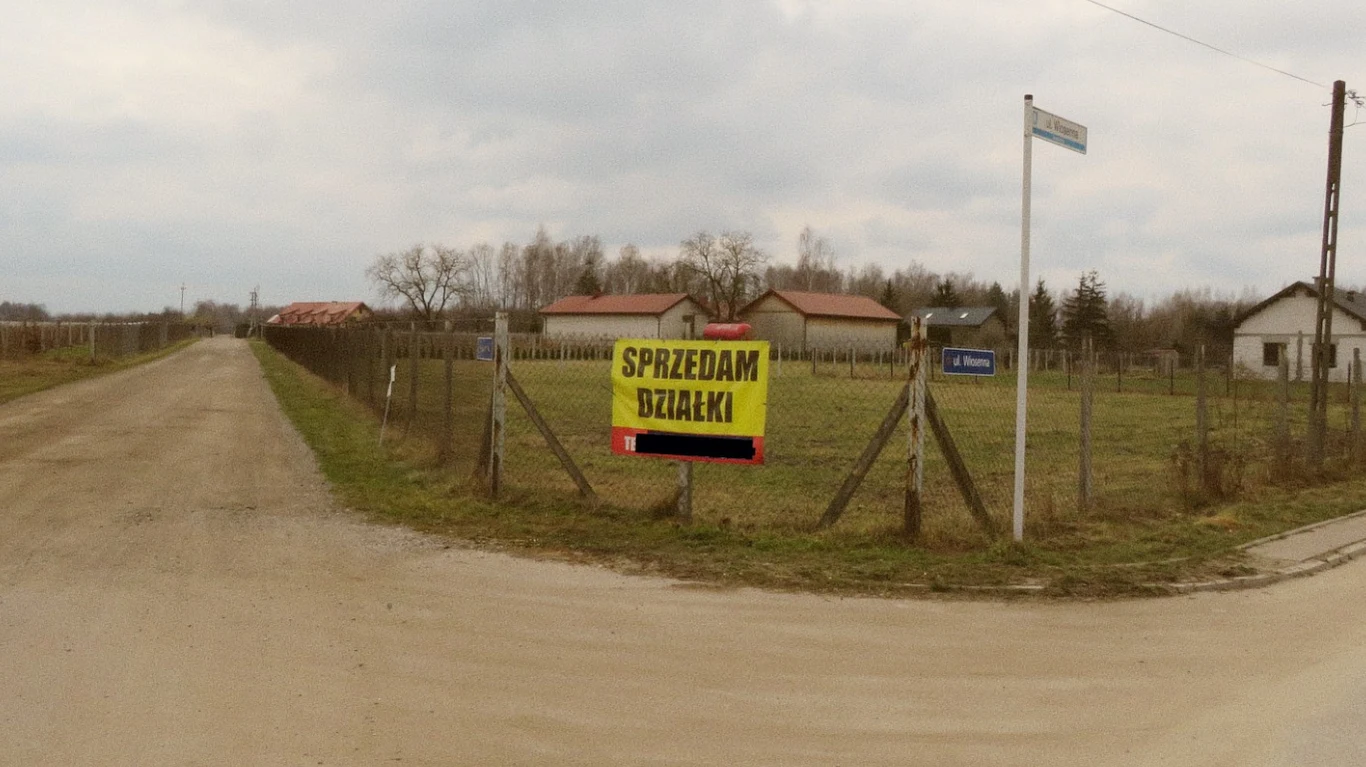 duża żółta tablica z napisem 'SPRZEDAM DZIAŁKI' umieszczona na ogrodzeniu działki przy skrzyżowaniu wiejskich dróg, w tle widoczne domy jednorodzinne oraz pola