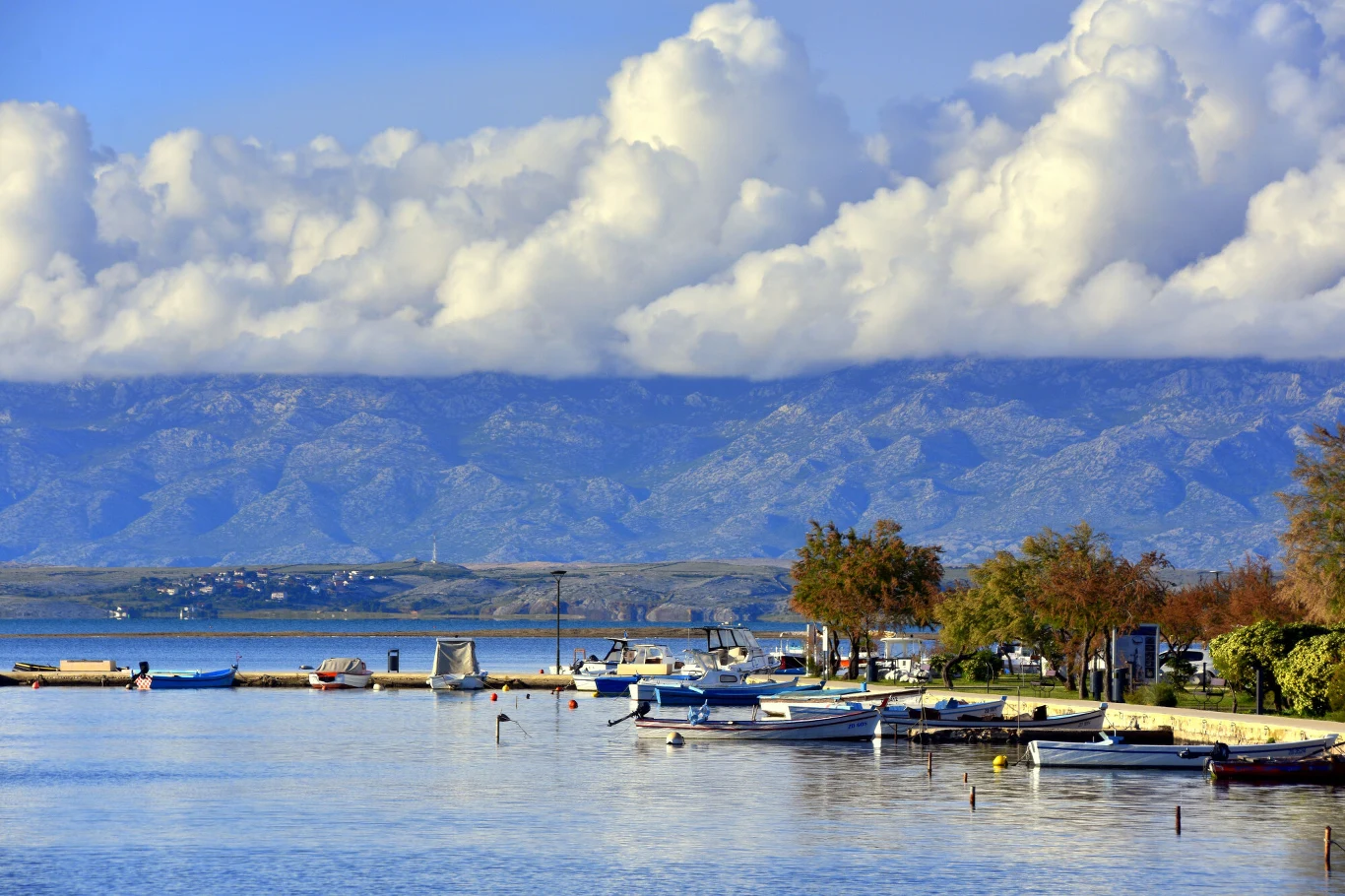 Mała marina z łodziami przycumowanymi do brzegu, spokojna woda odbijająca niebo, w tle monumentalne górskie pasmo częściowo zasłonięte przez gęste, białe chmury oraz zieleń i drzewa wzdłuż nabrzeża.
