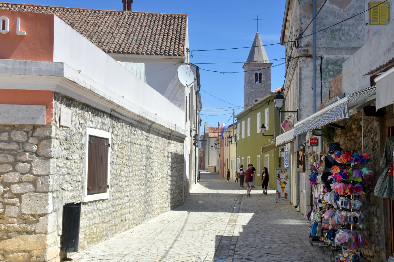 wąska brukowana uliczka z kamiennymi domami, po prawej stronie stragan z kolorowymi kapeluszami, w tle widoczna kamienna wieża kościoła z krzyżem na szczycie, kilka osób spaceruje w oddali