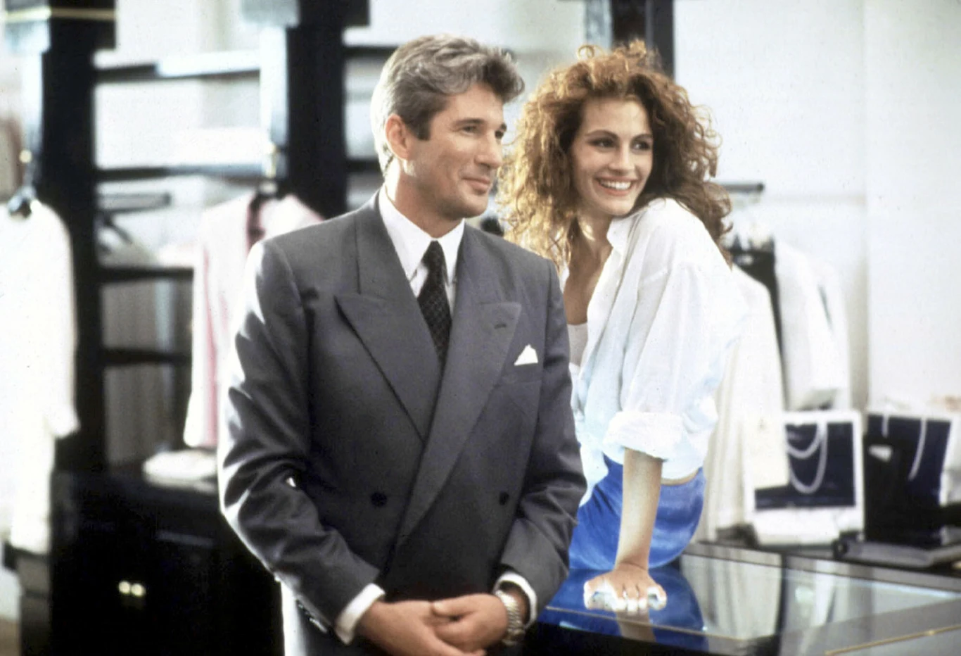 "Pretty Woman" mężczyzna w eleganckim szarym garniturze stoi obok uśmiechniętej kobiety z burzą rudych włosów, ubranej w białą bluzkę i niebieską spódnicę, oboje opierają się o szklaną ladę w jasnym, luksusowym butiku