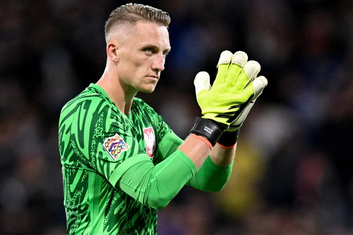 bramkarz w zielonym stroju sportowym z godłem Polski na piersi stoi z uniesionymi i złożonymi w geście braw dłoniami, patrząc przed siebie z poważnym wyrazem twarzy