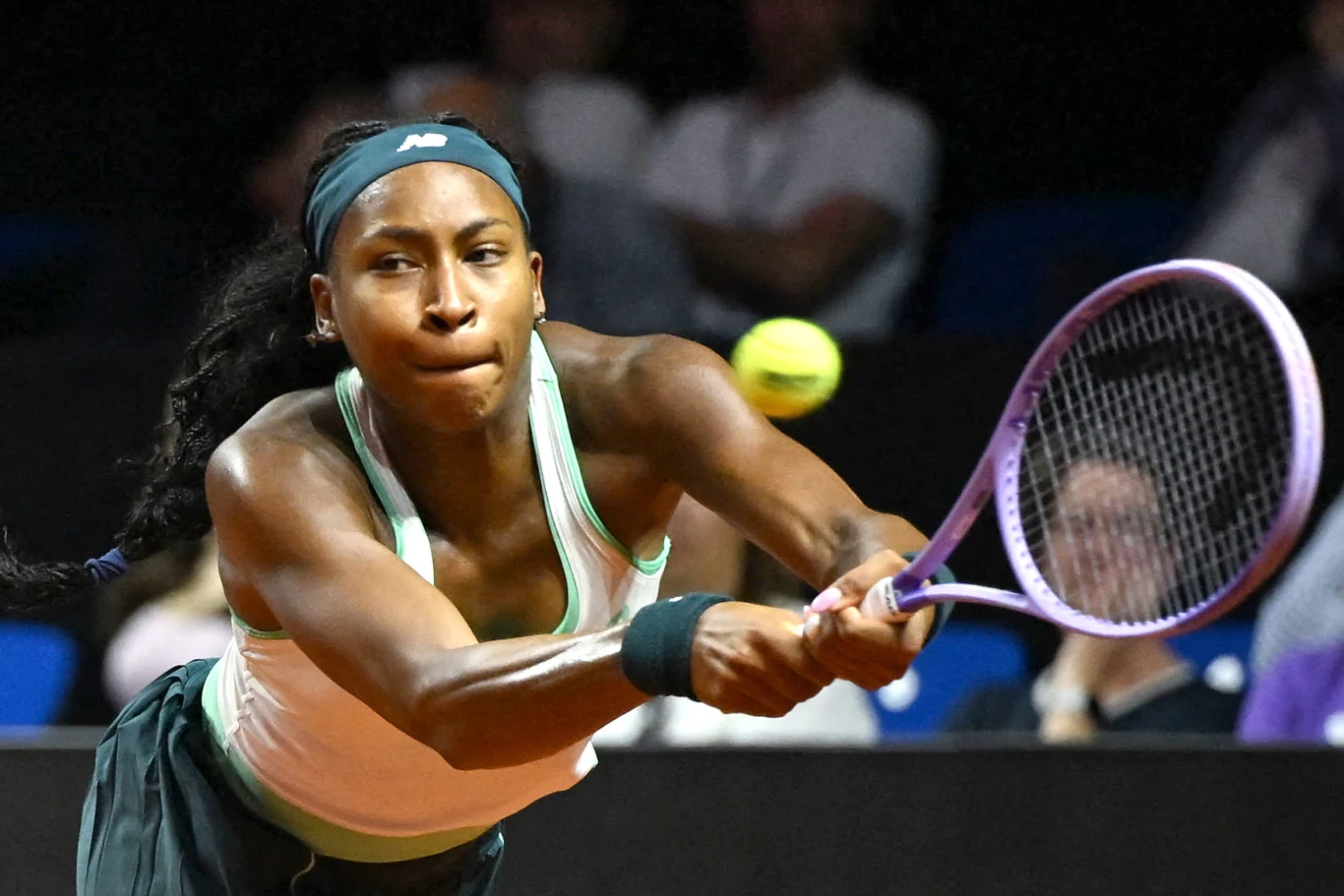 Kobieta w sportowym stroju grająca w tenisa, dynamicznie odbija piłkę rakietą, skoncentrowana i pełna determinacji, w tle widoczni nieostro kibice.