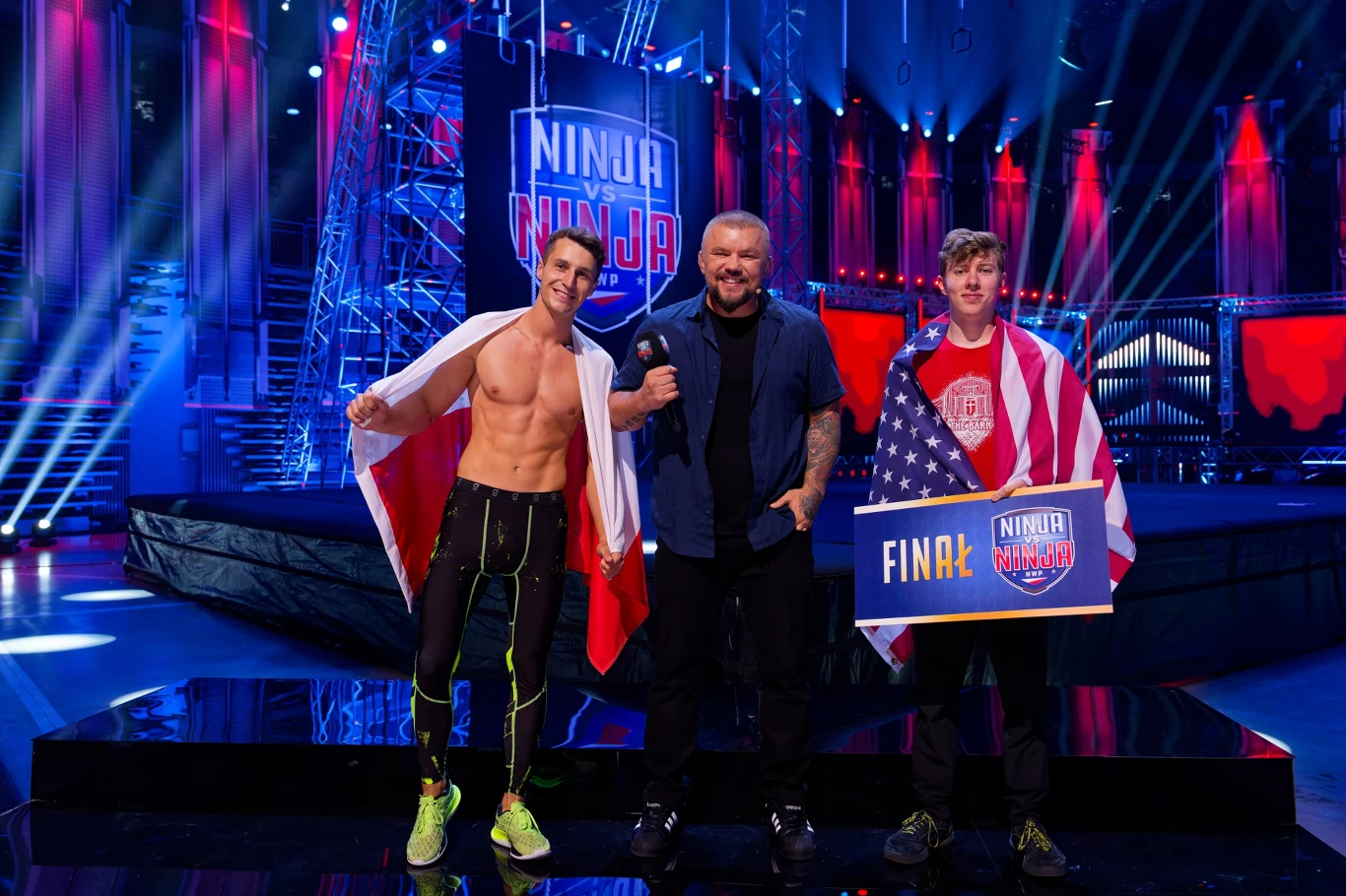 Damian Drzewiecki walczył o bezpośredni awans do finału z Samuelem Folsomem Dwóch młodych mężczyzn, jeden owinięty flagą Polski, drugi flagą Stanów Zjednoczonych, stoi po bokach uśmiechniętego prowadzącego na scenie sportowego show Ninja Warrior, otoczeni przez dynamiczne światła i futurystyczną scenografię.