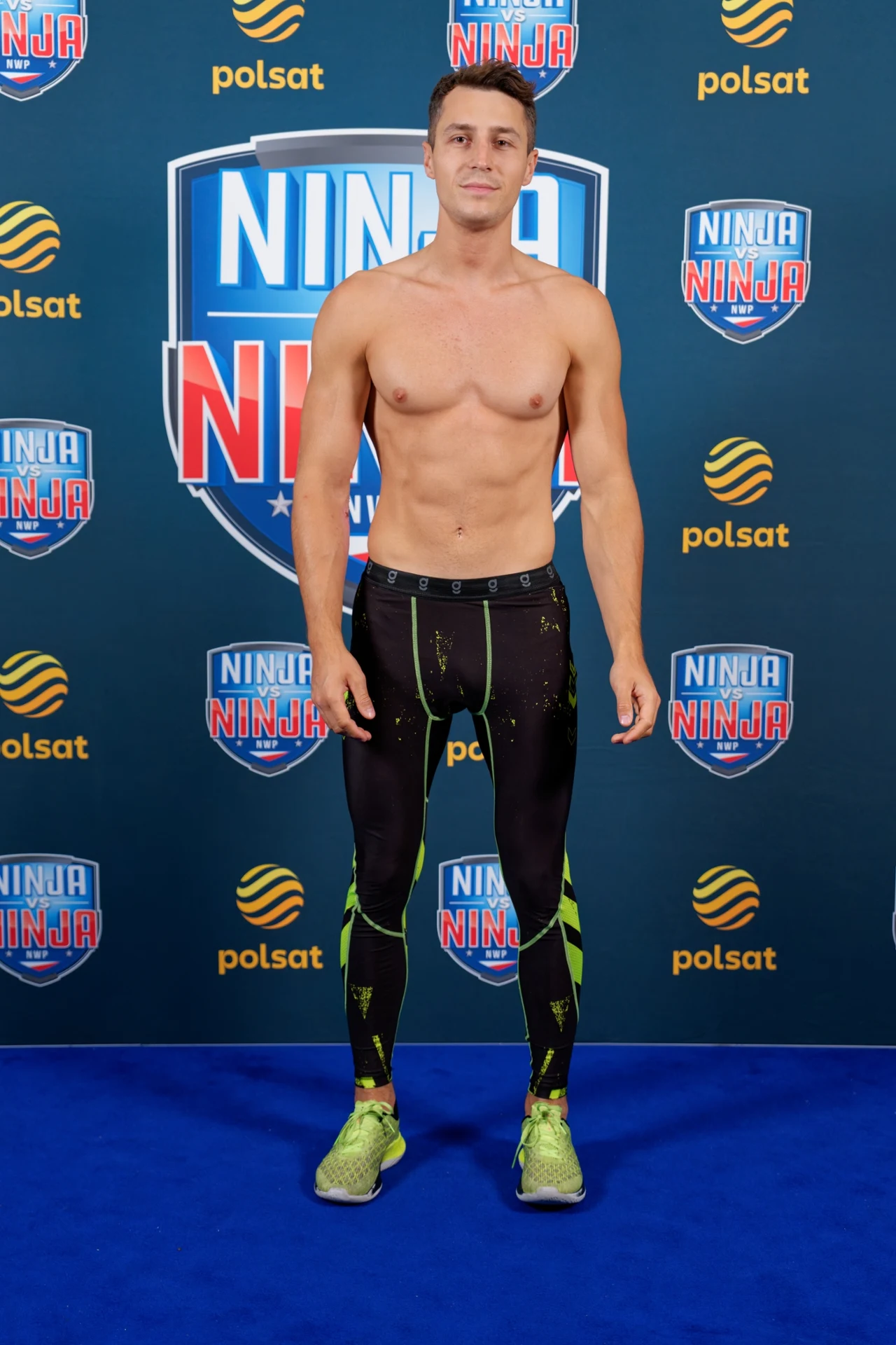 Damian Drzewiecki w Ninja vs Ninja Młody mężczyzna o wysportowanej sylwetce stoi na tle ścianki z logo programu Ninja Warrior Polska, ubrany w obcisłe legginsy sportowe z zielonymi akcentami i neonowe buty sportowe, bez koszulki.