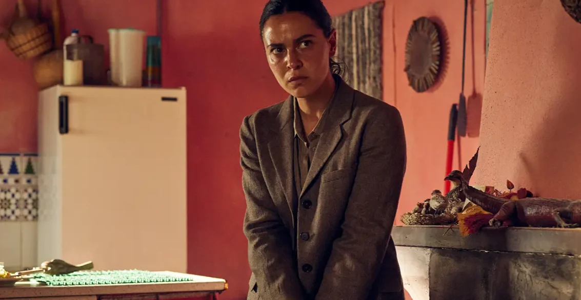 Kadr z filmu "Mumia: Film Lee Cronina" Kobieta w garniturze stoi w kuchni o różowych ścianach przy lodówce i stole z zielonym obrusem.