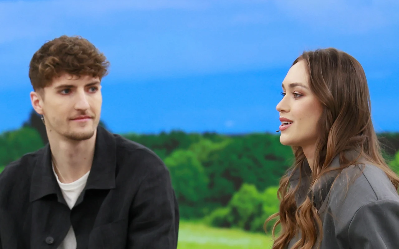 Daria Syta z narzeczonym w "halo tu polsat" Dwoje młodych ludzi w casualowych ubraniach siedzi naprzeciw siebie, prowadząc rozmowę na tle krajobrazu z zielonymi drzewami i niebieskim niebem.