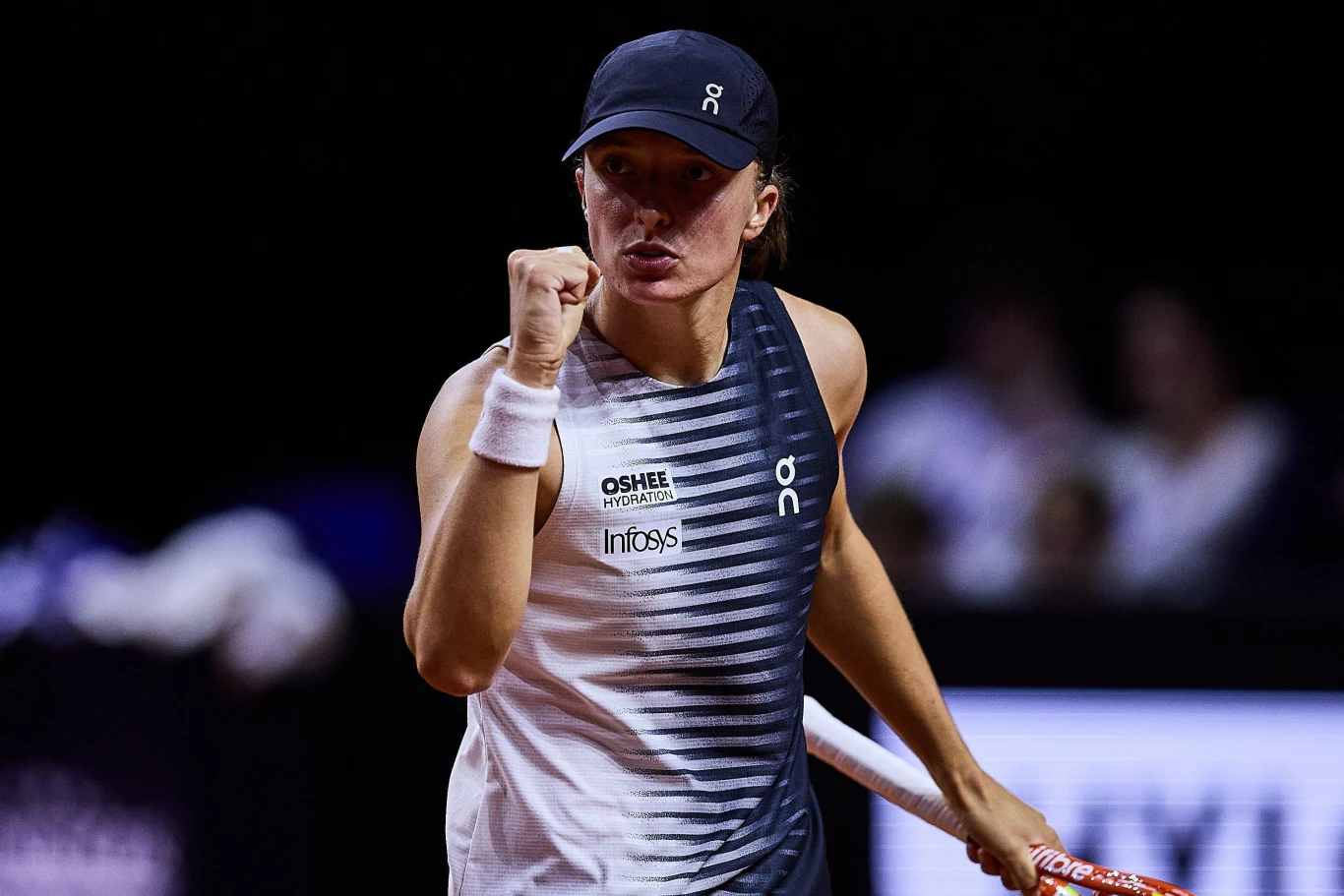 Tenisistka w sportowej odzieży i czapce, z zaciśniętą pięścią w geście triumfu, trzyma rakietę i patrzy skupionym wzrokiem w kierunku przeciwnika.