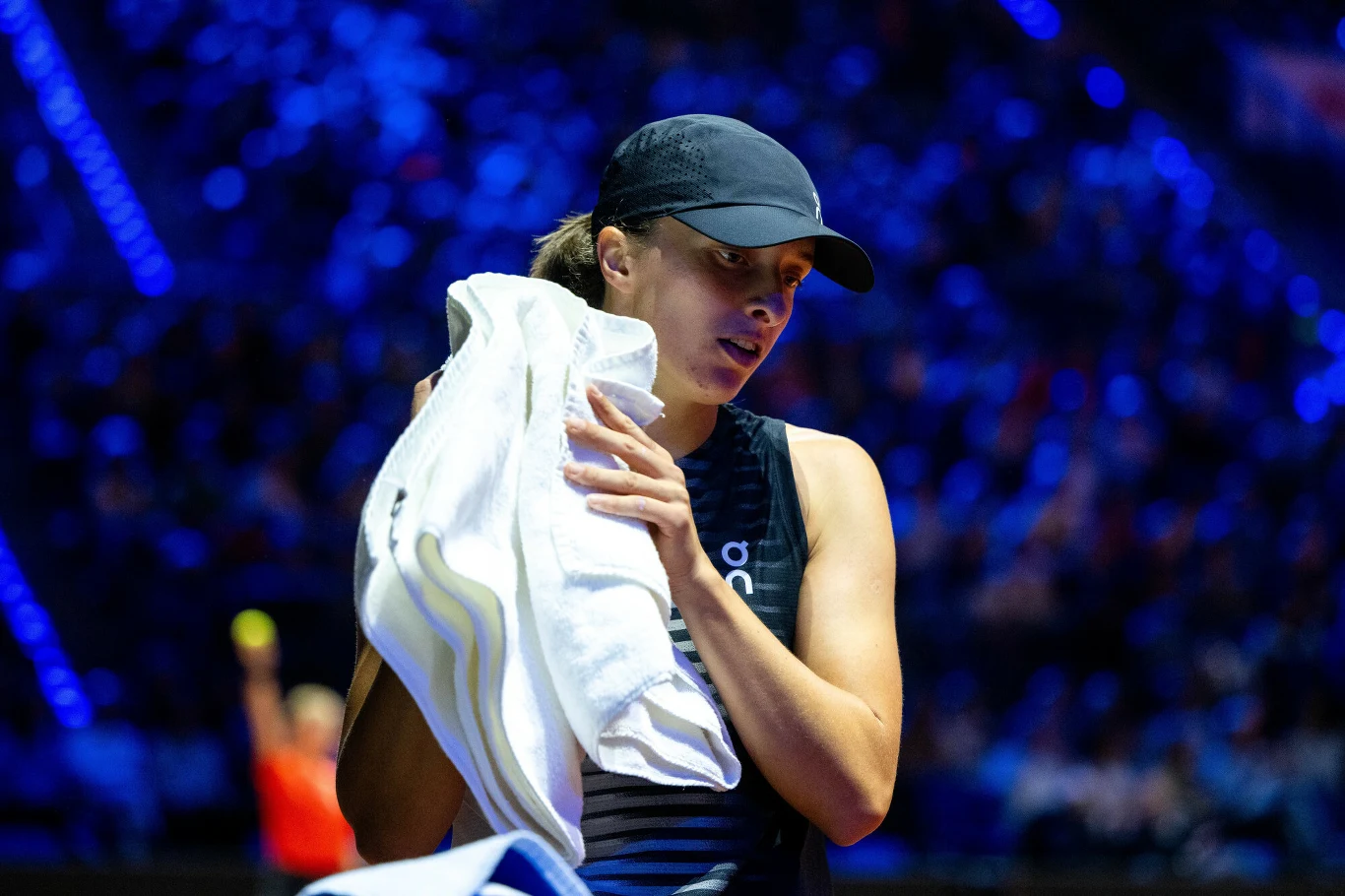 tenisistka w czapce z daszkiem trzymająca biały ręcznik, ubrana w sportową odzież, stoi na tle rozświetlonej niebieskimi światłami widowni