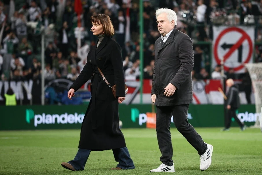 dwie osoby w eleganckich płaszczach idą po murawie stadionu piłkarskiego, w tle tłum kibiców i duży transparent z przekreślonym symbolem