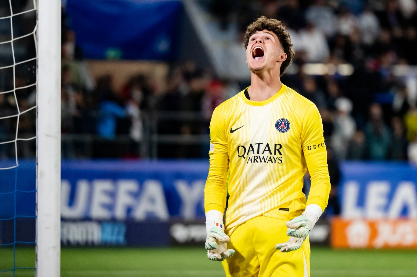 Real Madryt - PSG w młodzieżowej LM bramkarz w żółtym stroju z logo PSG, stojący przed bramką z uniesioną głową i szeroko otwartymi ustami, wyrażający silne emocje; w tle rozmyta publiczność na trybunach oraz baner z napisem UEFA