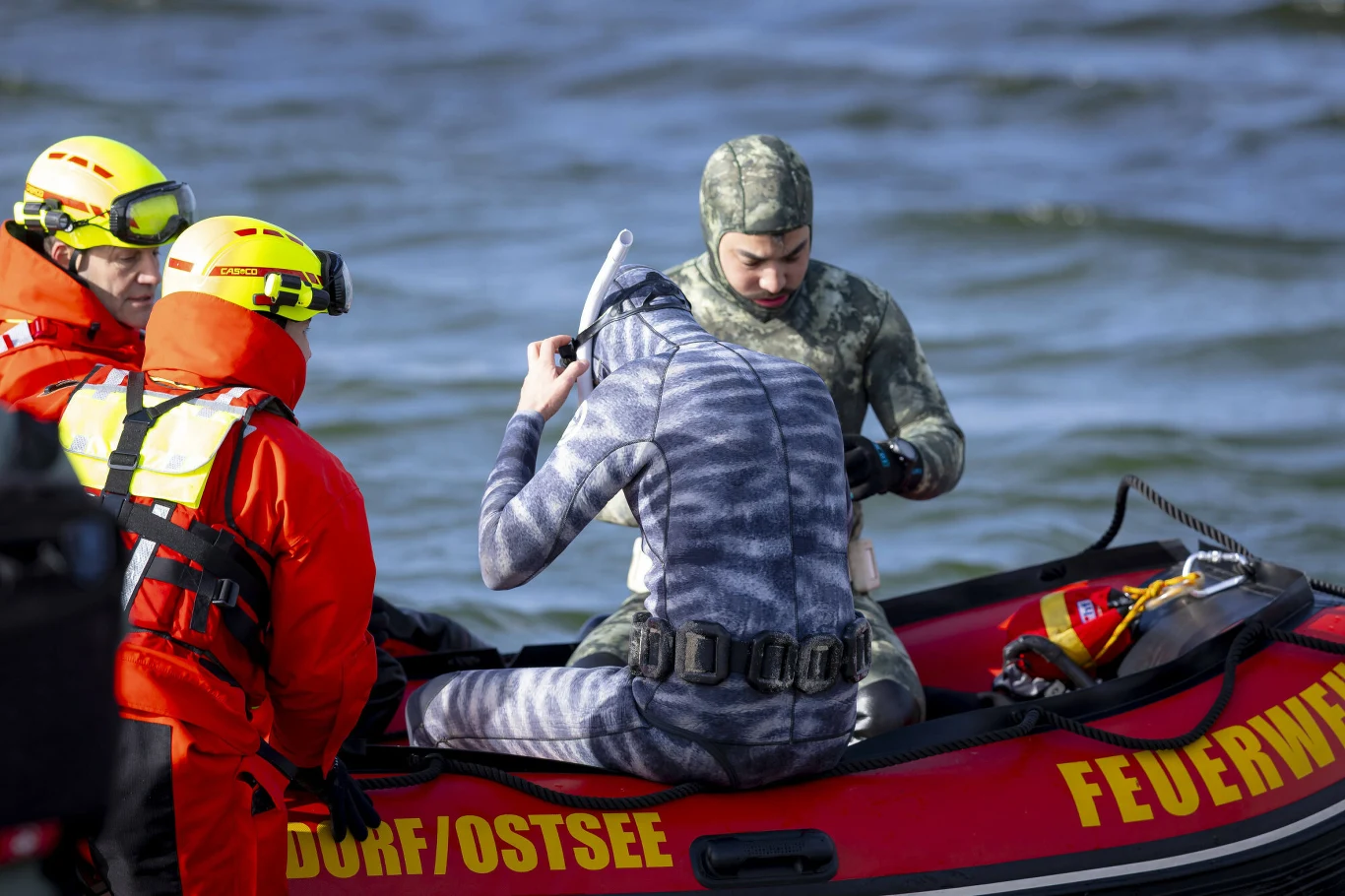 Dwóch ratowników w pomarańczowych ubraniach ochronnych stoi na pontonie oznaczonym napisem 'FEUERWEHR', obok dwóch nurków w piankach i maskach, z których jeden poprawia sprzęt do nurkowania; tło stanowi wzburzona woda.