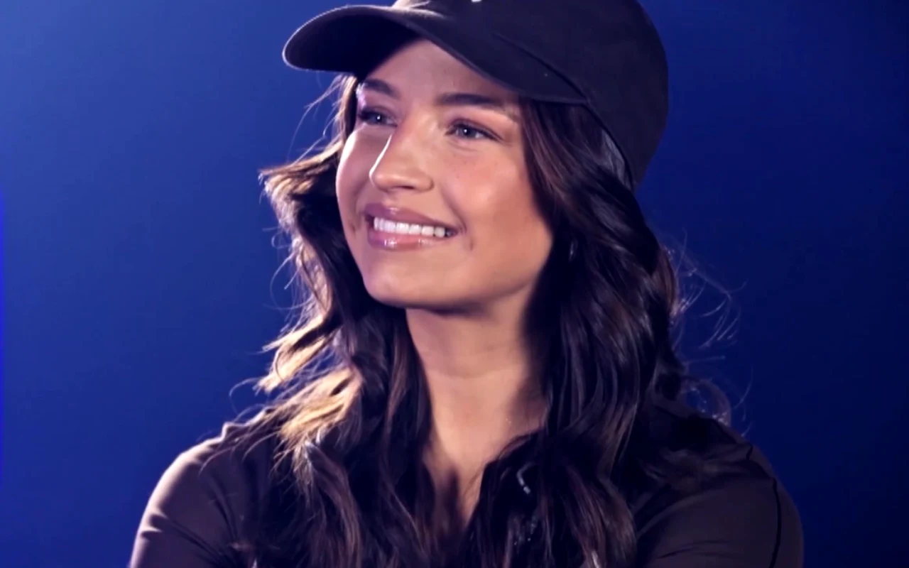 Młoda kobieta o długich, ciemnych włosach z delikatnym makijażem, uśmiechnięta, ubrana w czapkę z daszkiem i ciemną bluzę, na tle jednolitego, ciemnoniebieskiego tła.