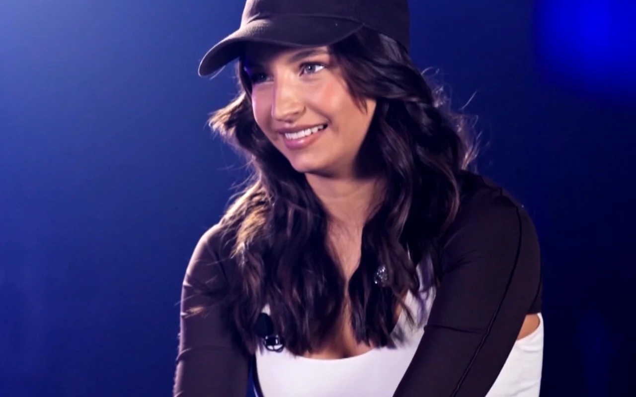 Młoda kobieta z długimi, ciemnymi, falującymi włosami uśmiecha się, ubrana w czapkę z daszkiem oraz biało-czarną bluzę, na tle ciemnoniebieskiego rozmytego tła.