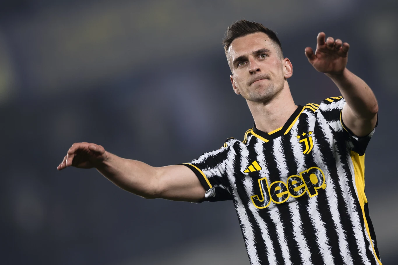 Arkadiusz Milik Arkadiusz Milik w biało-czarnej koszulce z logo Jeep z uniesionymi rękoma na tle rozmytego stadionu