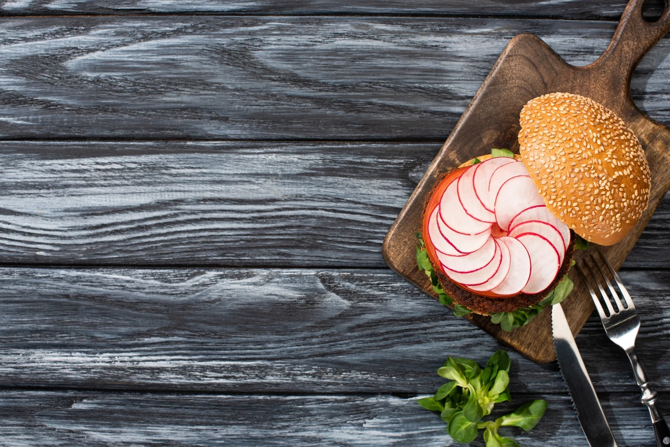top view of tasty vegan burger with microgreens, radish, tomato on cutting board with cutlery served on wooden table