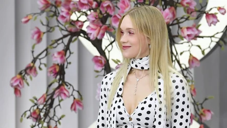 kobieta w długich blond włosach ubrana w elegancką białą bluzkę w czarne grochy, ozdobiony perłowy naszyjnik, w tle gałęzie drzewa magnolii z kwitnącymi różowymi kwiatami