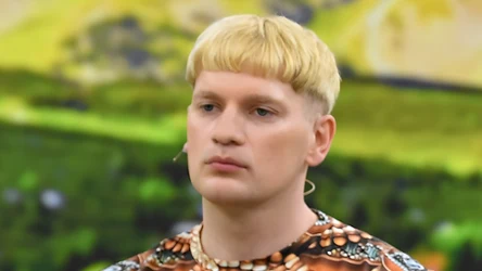 Młody mężczyzna o jasnych blond włosach z prostą grzywką, ubrany w koszulkę z kolorowym, geometrycznym wzorem i koralikami wokół szyi, stoi na tle rozmytego, zielonego krajobrazu.