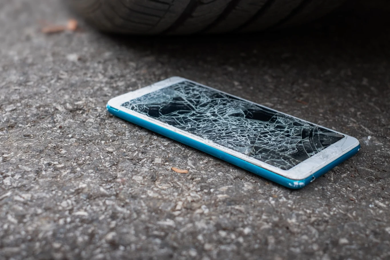 Do naprawy smartfonów coraz częściej wykorzystuje się techniki znane z rynku motoryzacyjnego Smartfon z rozbitym ekranem na asfalcie obok opony samochodu, przed naprawą techniką motoryzacyjną