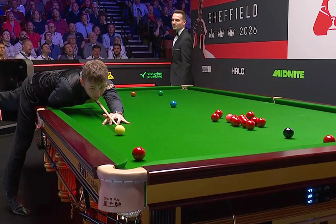 zawodnik w trakcie uderzenia snookerowego przy stole bilardowym, liczne kule ustawione na zielonym suknie, sędzia stojący obok stołu, w tle widownia i baner z napisem SHEFFIELD 2026