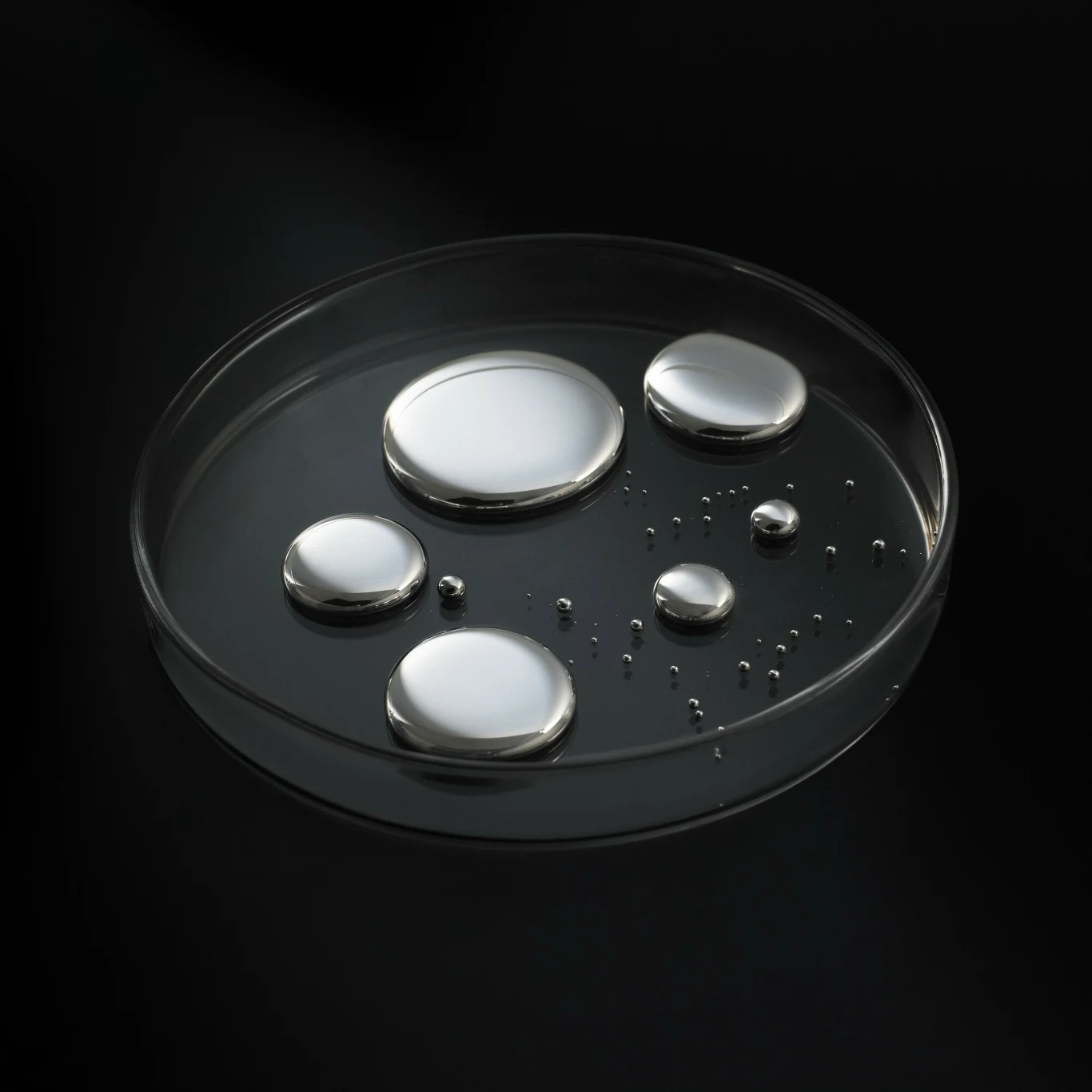 kilka dużych i małych kropli ciekłego srebrzystego metalu umieszczonych na szklanej szalce Petriego na czarnym tle