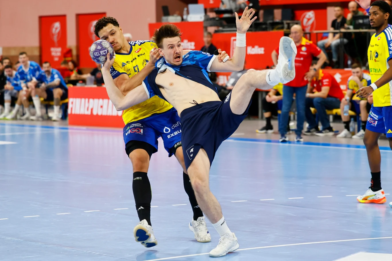dwóch szczypiornistów w dynamicznej walce o piłkę podczas meczu, jeden zawodnik w żółto-niebieskim stroju blokuje przeciwnika w niebieskiej koszulce, który traci równowagę i jest niemalże w powietrzu