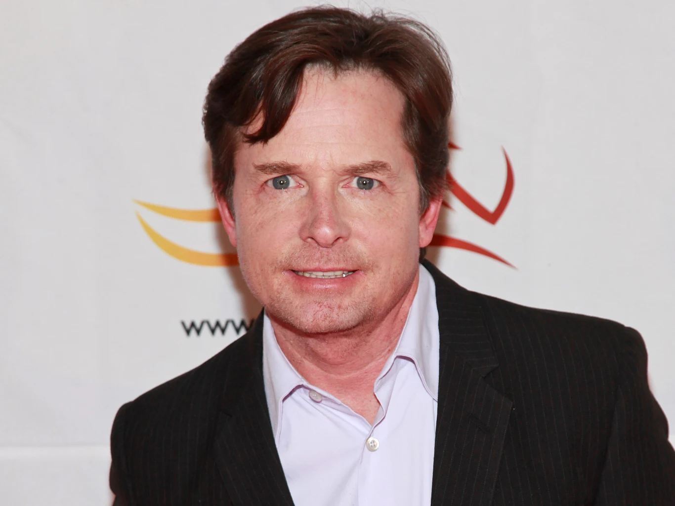 Michael J. Fox Mężczyzna z krótkimi brązowymi włosami w ciemnej marynarce i jasnej koszuli na tle jasnej ścianki z abstrakcyjnym wzorem.