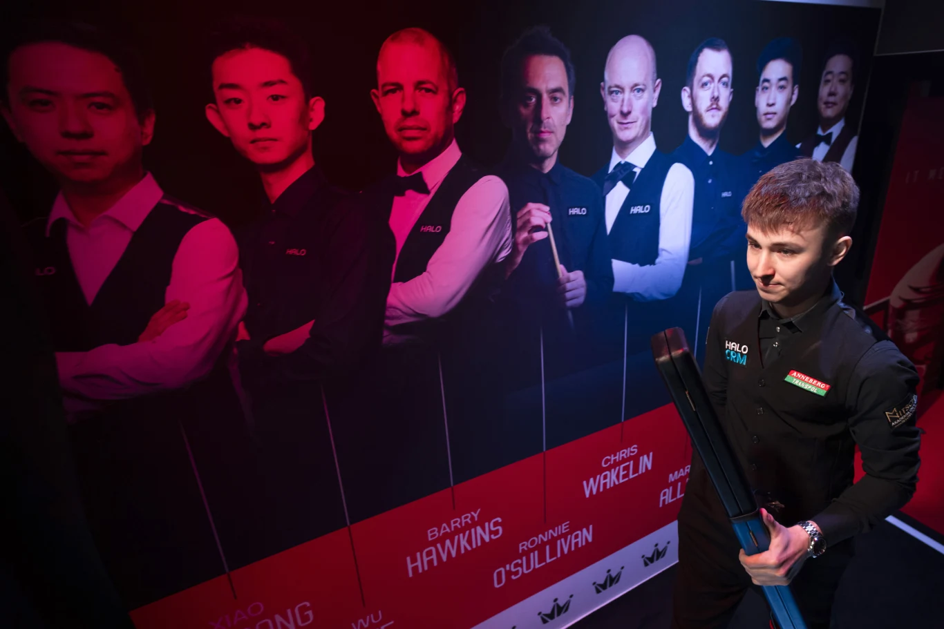 Młody snookerzysta w profesjonalnym stroju stoi obok dużego plakatu z wizerunkami znanych zawodników snookera, trzymając kij bilardowy i skupiony patrząc w bok.