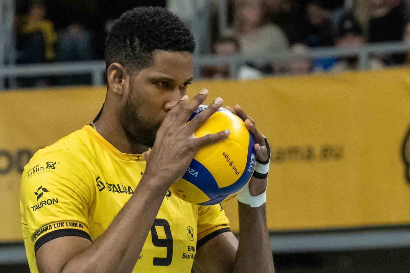 Wilfredo Leon, przyjmujący Bogdanki LUK Lublin siatkarz w żółtej koszulce z numerem 9 koncentruje się przed wykonaniem zagrywki, trzymając piłkę do siatkówki przy twarzy, w tle rozmyta publiczność i żółty baner