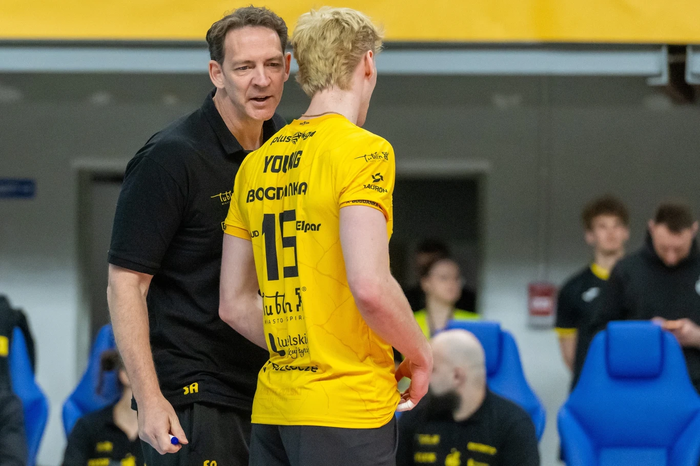 Stephane Antiga i Jackson Ypung trener w czarnej koszulce rozmawia z siatkarzem w żółtym stroju z numerem 15, w tle kilku członków drużyny i niebieskie krzesełka