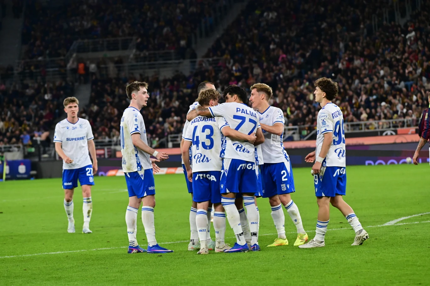 Piłkarze Lecha cieszą się z gola na 1:0 w Szczecinie Grupa piłkarzy drużyny w białych koszulkach z niebieskimi akcentami świętuje zdobycie gola na boisku, otoczona przez pełne trybuny kibiców.