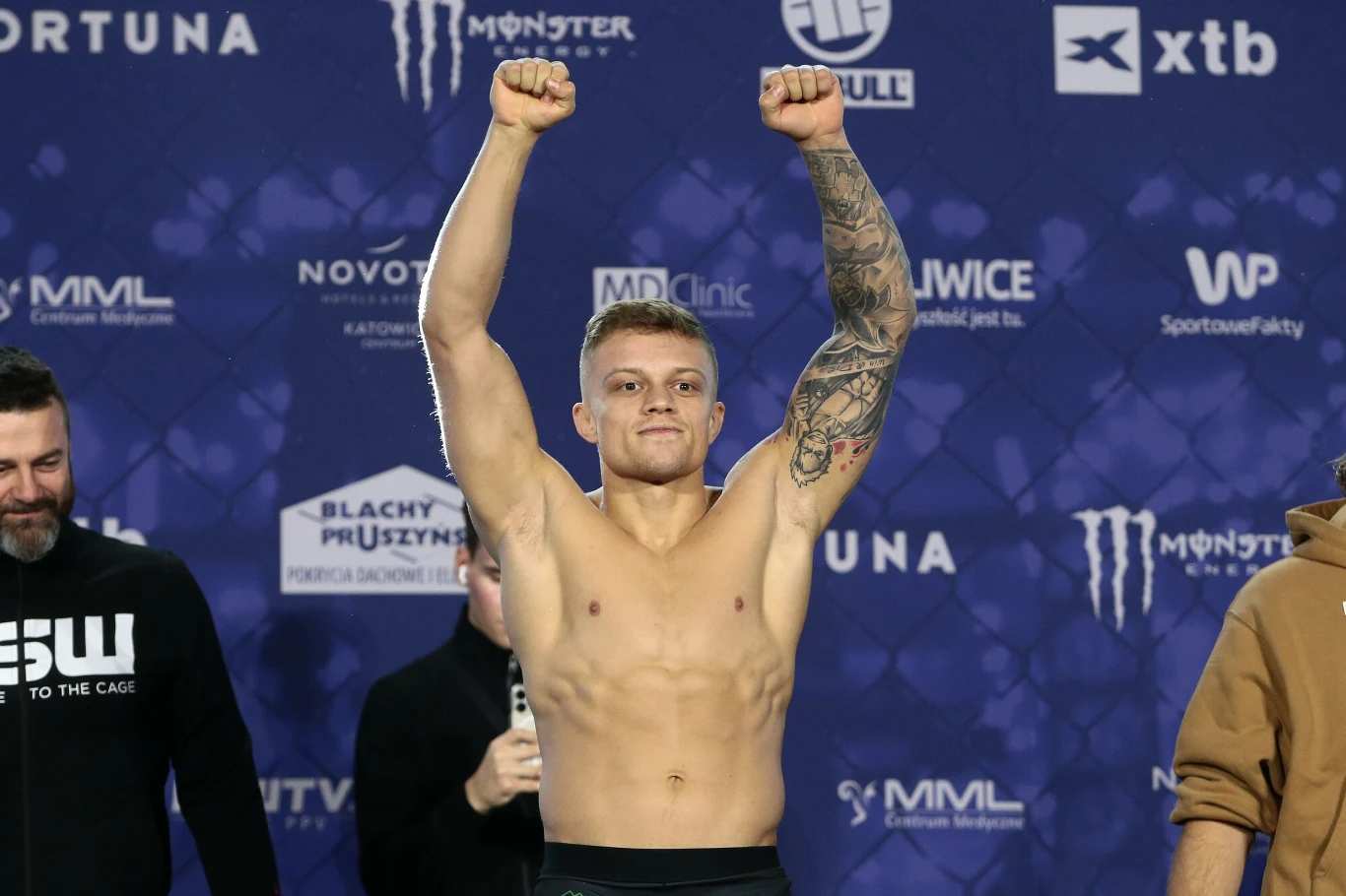 Patryk Kaczmarczyk umięśniony mężczyzna z uniesionymi rękami w geście zwycięstwa, tatuaż na prawym ramieniu, stoi na tle ściany z logotypami sponsorów