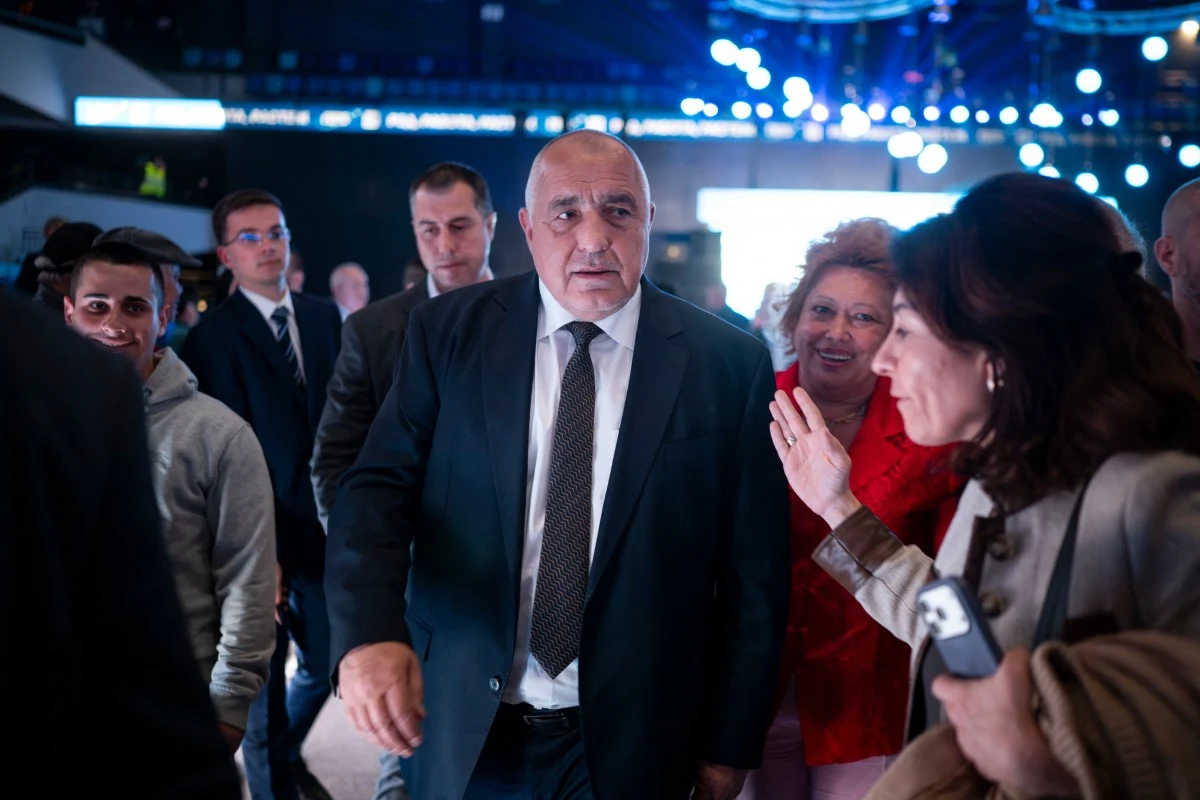 Bojko Borisow, były premier i lider partii GERB mężczyzna w garniturze stoi w centrum grupy ludzi podczas wydarzenia w oświetlonym wnętrzu, wokół niego znajdują się osoby w eleganckich i codziennych ubraniach, tło rozświetlone jest licznymi punktami świetlnymi