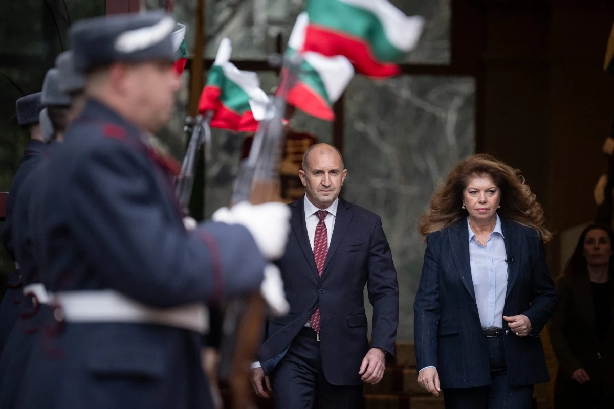 Mężczyzna i kobieta w eleganckich garniturach idą obok siebie, patrząc poważnie przed siebie; po obu stronach stoją mundurowi żołnierze trzymający flagi Bułgarii.