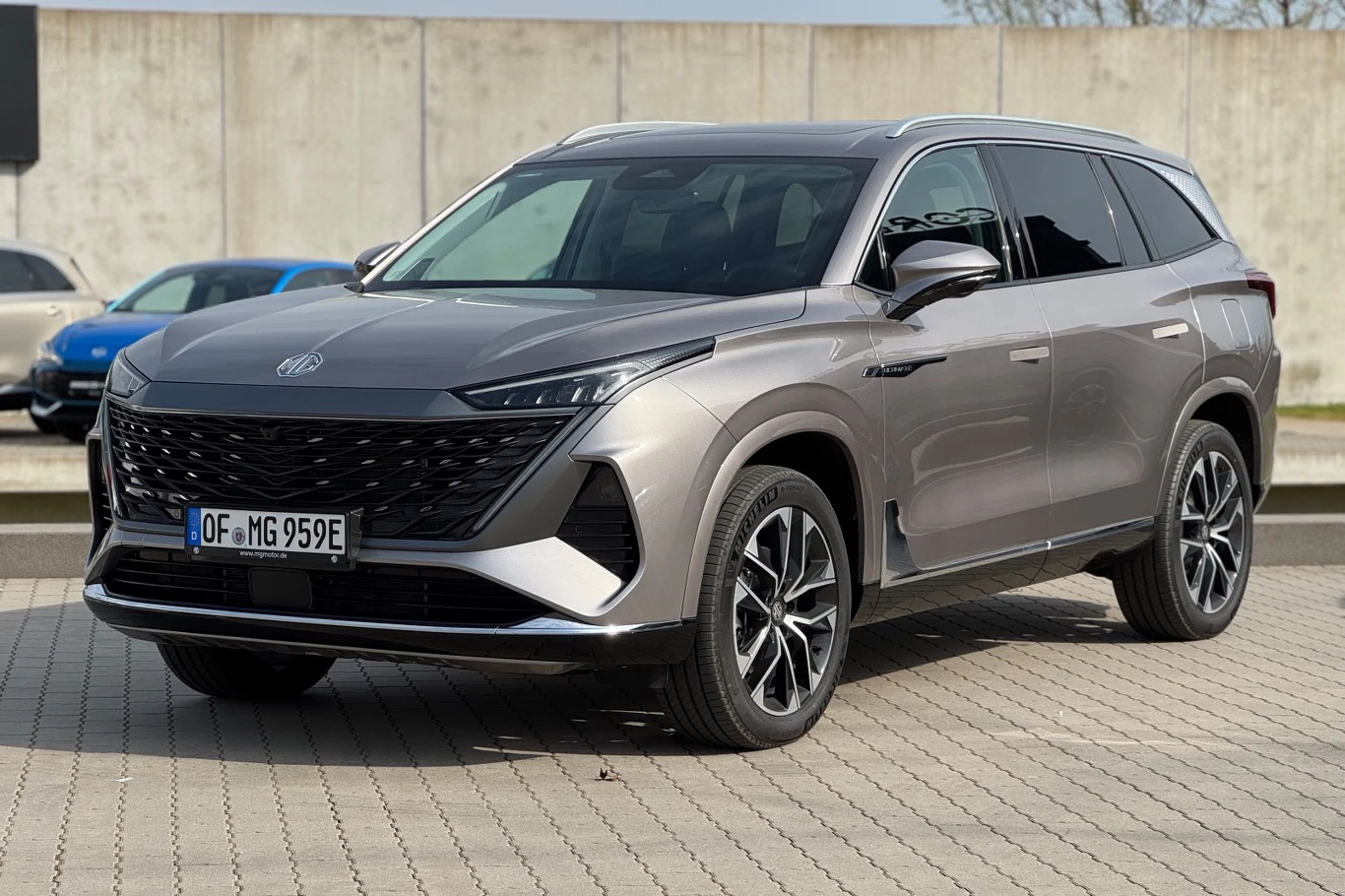 MG S9 kusi klientów, którzy szukają 7-miejscowego SUV-a MG S9, srebrnoszary 7-miejscowy SUV plug-in hybrid, zaparkowany na brukowanym placu przy betonowym murze