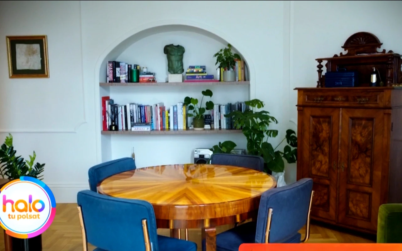 okrągły drewniany stół otoczony niebieskimi krzesłami, w tle półki z książkami i roślinami umieszczone we wnęce ściennej, po prawej stronie zabytkowa komoda, na podłodze parkiet