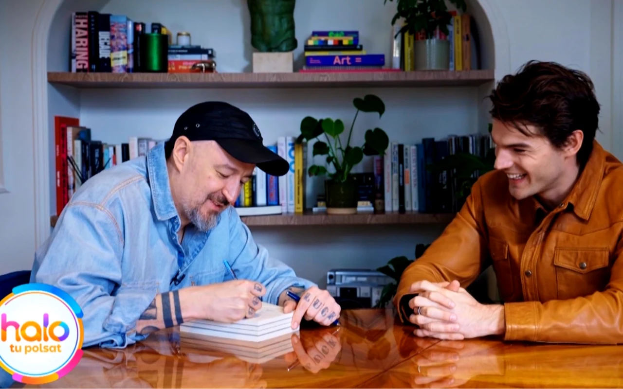 Dwóch mężczyzn siedzi przy drewnianym stole, jeden z nich podpisuje stos książek, drugi się uśmiecha; w tle półka z książkami i rośliną doniczkową, w lewym dolnym rogu kolorowe logo „halo tu polsat”.