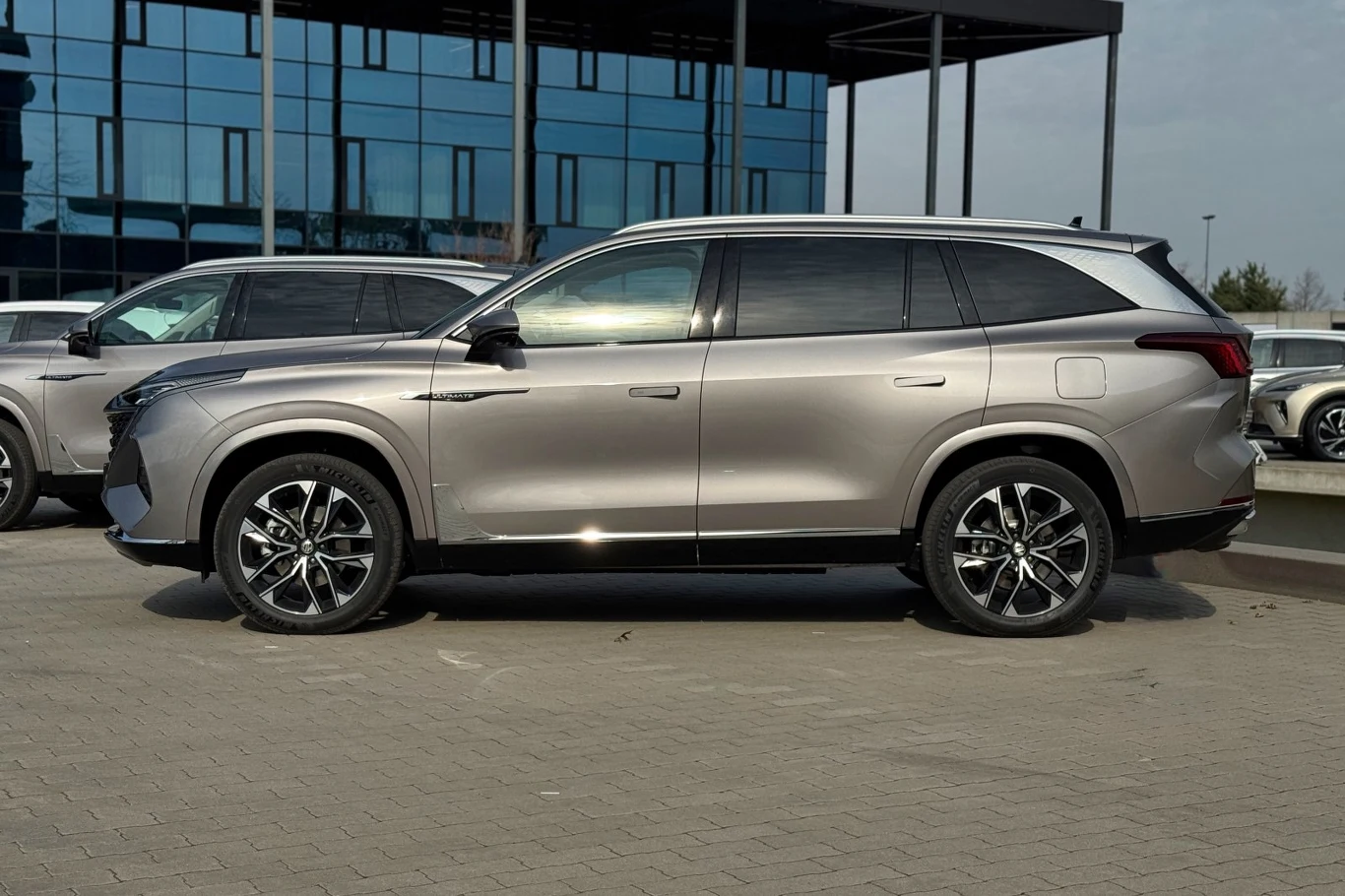 Nadwozie MG S9 ma 4983 mm długości, a rozstaw osi wynosi 2915 mm srebrny SUV o nowoczesnej sylwetce z czarnymi akcentami na felgach i linii okien, zaparkowany na utwardzonym placu przy szklanym budynku