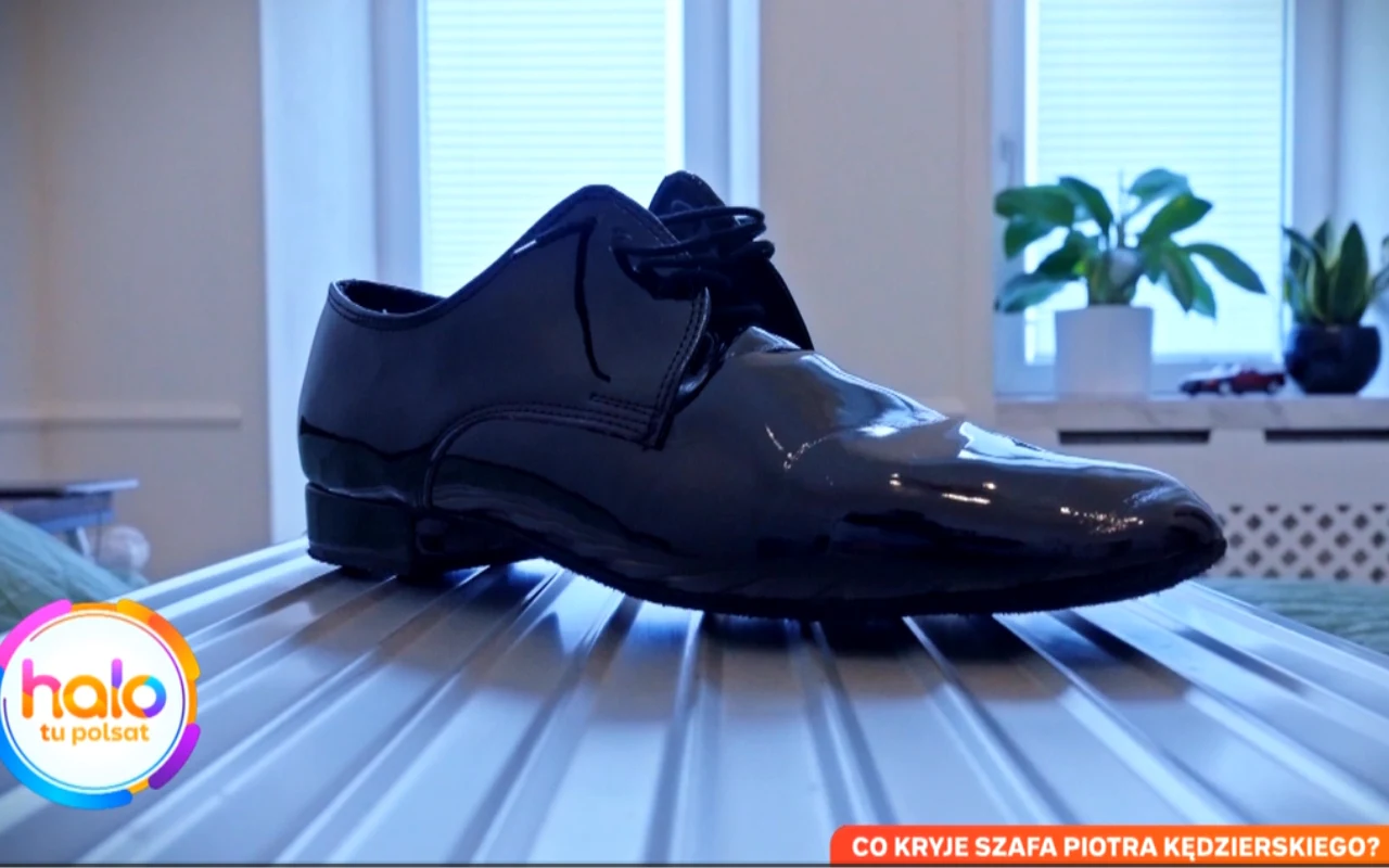 czarny lakierowany but męski ustawiony na srebrnej walizce w jasnym, nowocześnie urządzonym pokoju, w tle rośliny doniczkowe na parapecie i zamknięte żaluzje na oknach